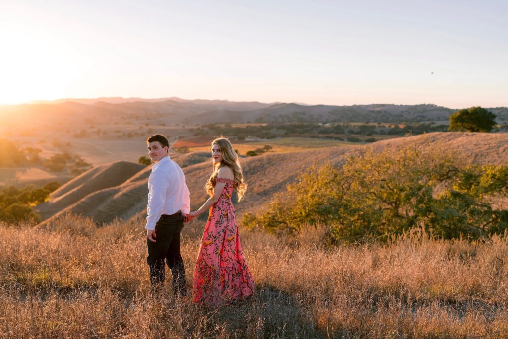 Woman in pink dress holding fiancee's hand in the hills at sunset