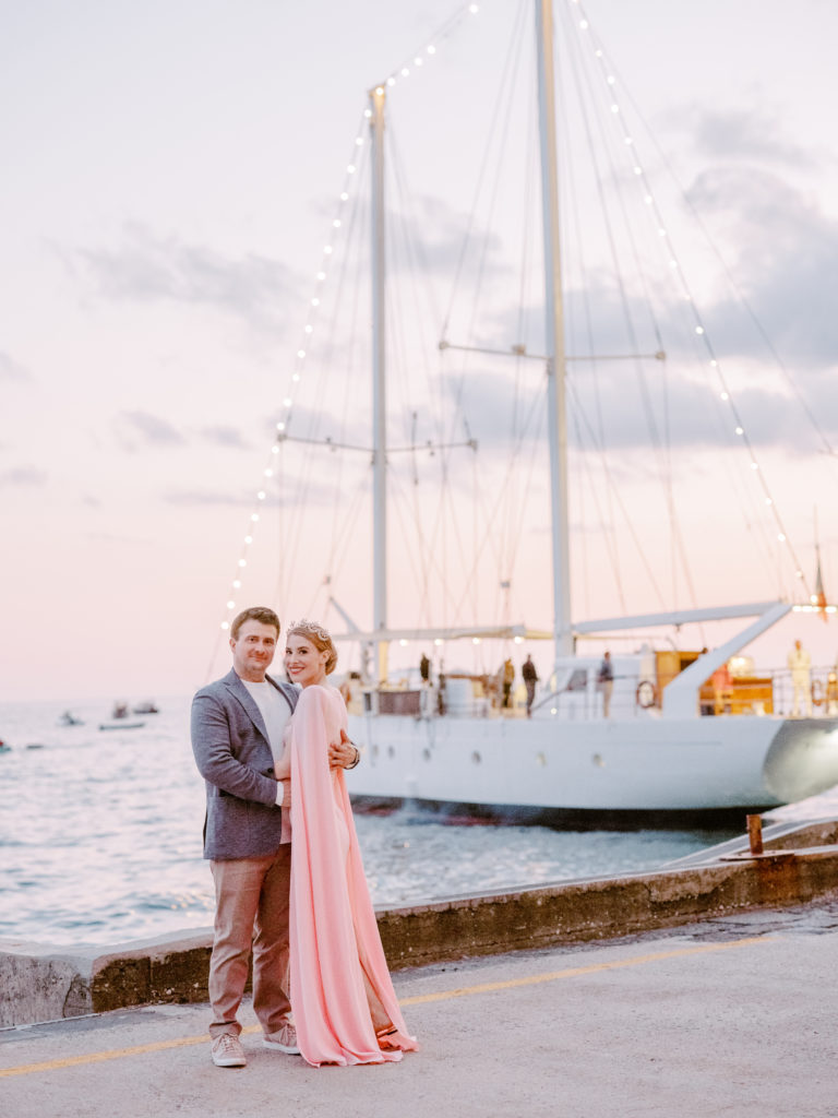Couple standing in front of a sailboat at sunset.