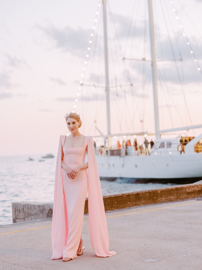 Woman in a pink evening dress standing in front of a sailboat docked in the Amalfi Coast.